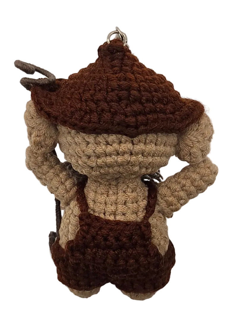 The back of a crocheted character toy, wearing a brown cap with brown overalls, lying on a plain white surface.