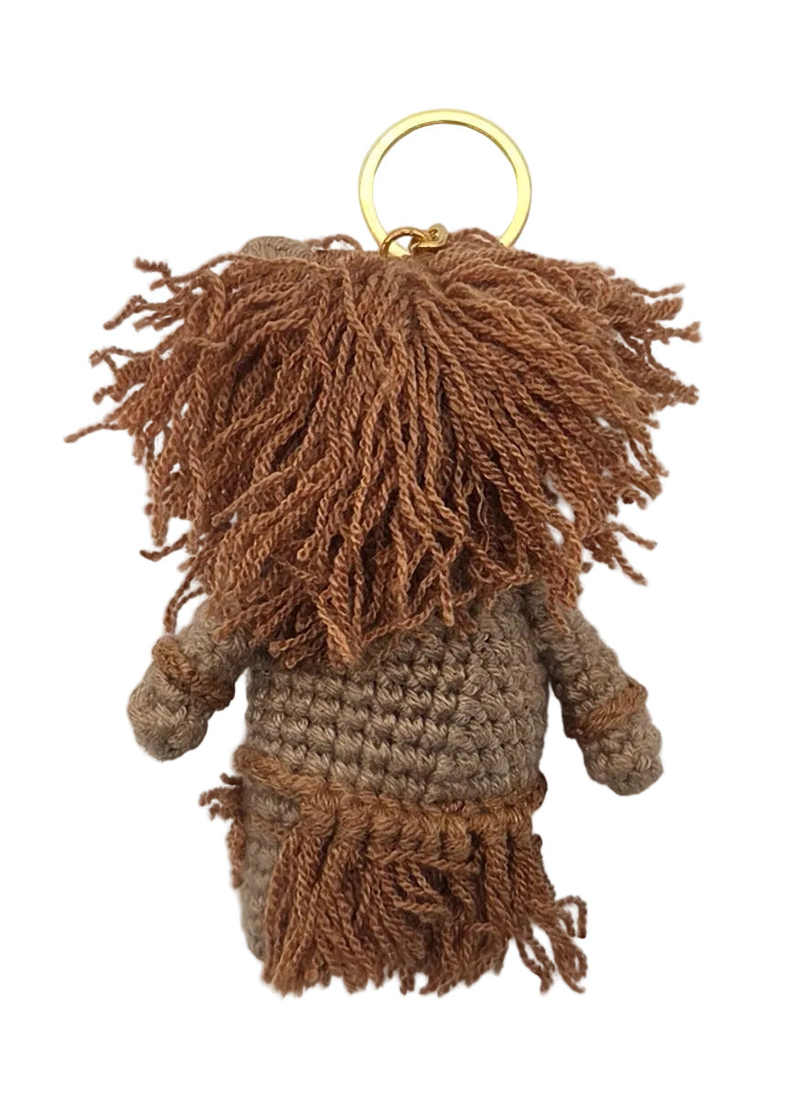 The back of a crochet keychain with brown yarn hair, a beige body, and a gold keychain at the top, shown on a white surface.