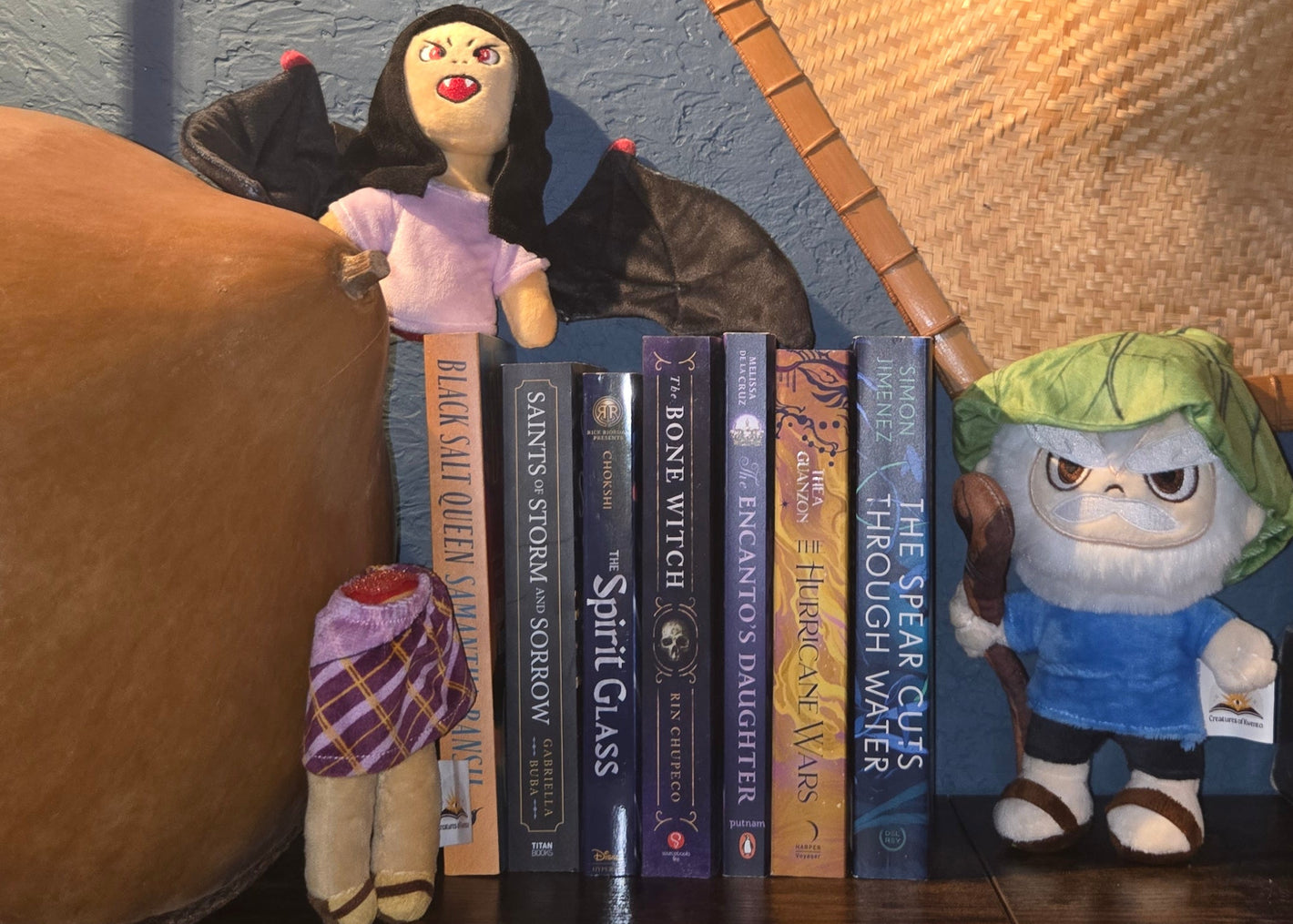Line of books on a surface between a gourd hat and dwende plushie and a manananggal plushie on top