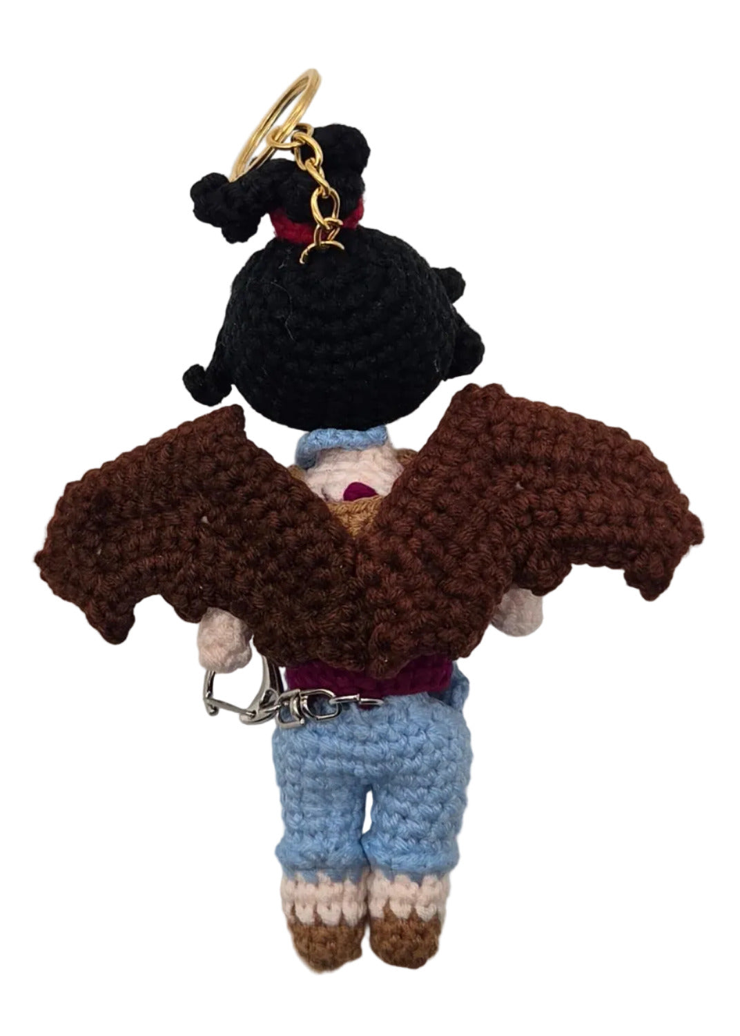 The back of a crocheted doll on a white background. The figure has black hair, brown wings, and light blue pants, with a small gold keychain attached.