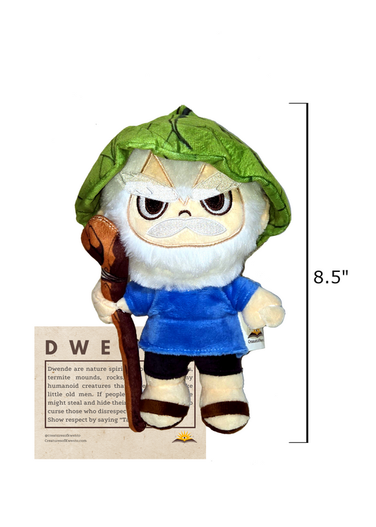 A dwende plush. It is a grumpy looking man with pale skin, white hair and beard, thick white eyebrows, and white mustache. He is wearing a green hat that looks like a leaf, a blue shirt, and black pants. In his right hand, he is holding what looks like a wood cane. On the side of the plush is a line marking the height of it as 8.5". The plush is over a brown summary sheet of the Dwende.