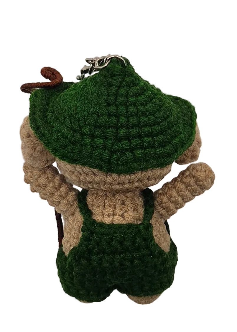 The back of a crocheted character toy, wearing a green cap with green overalls, lying on a plain white surface.