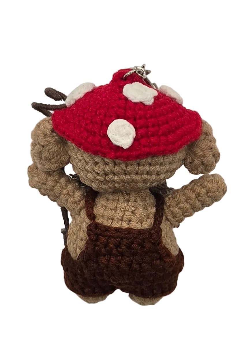 The back of a crocheted character toy, wearing a mushroom cap with brown overalls, lying on a plain white surface.